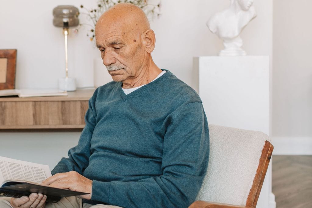 A white-haired man reading a book without glasses, sitting in an armchair in a light-walled room.