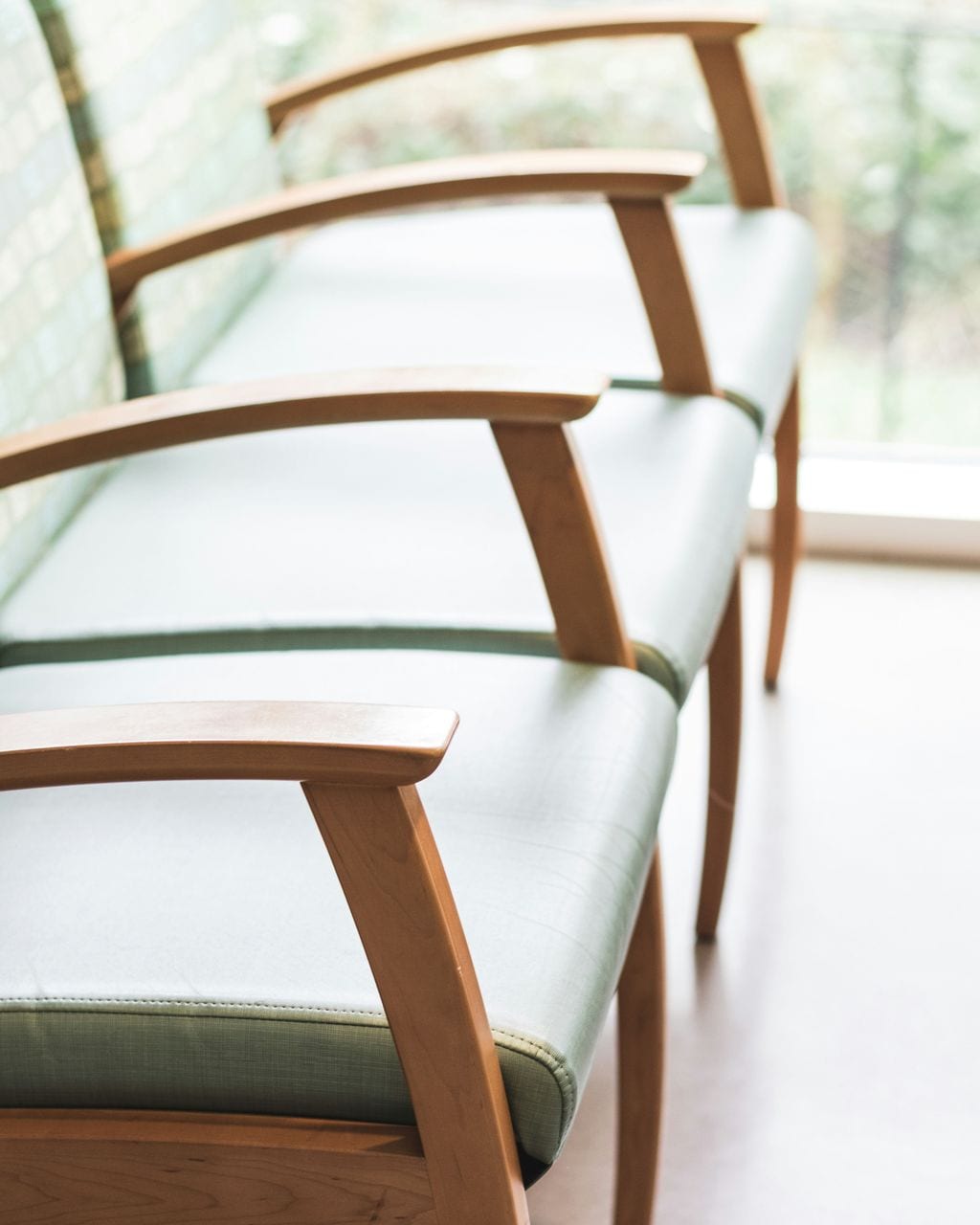 A row of wooden chairs with light green seats in a waiting room lit by a window.