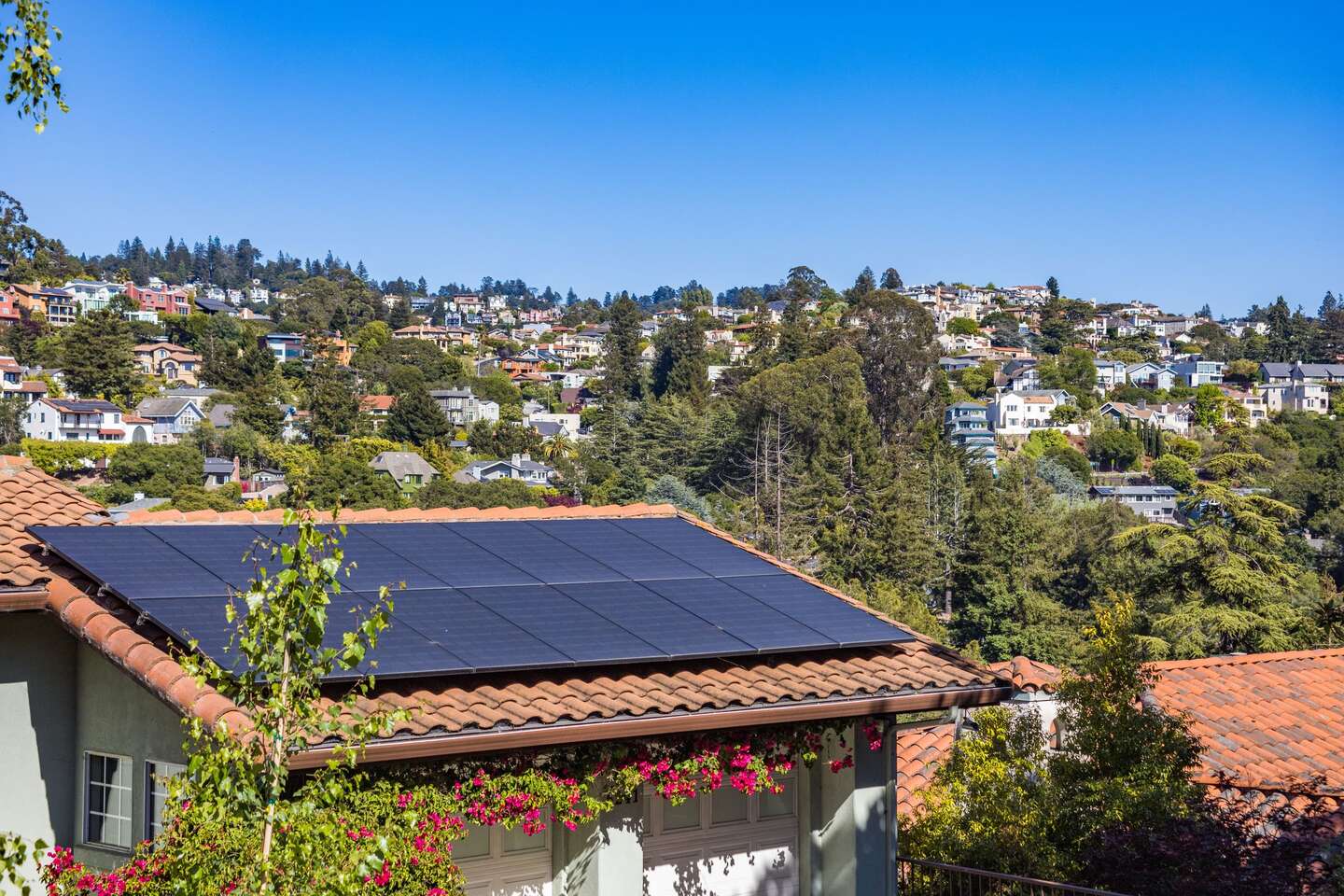 House with a clay-tile roof and a sixteen-panel solar array on the plane facing the sun, with a hillside neighborhood behind it.