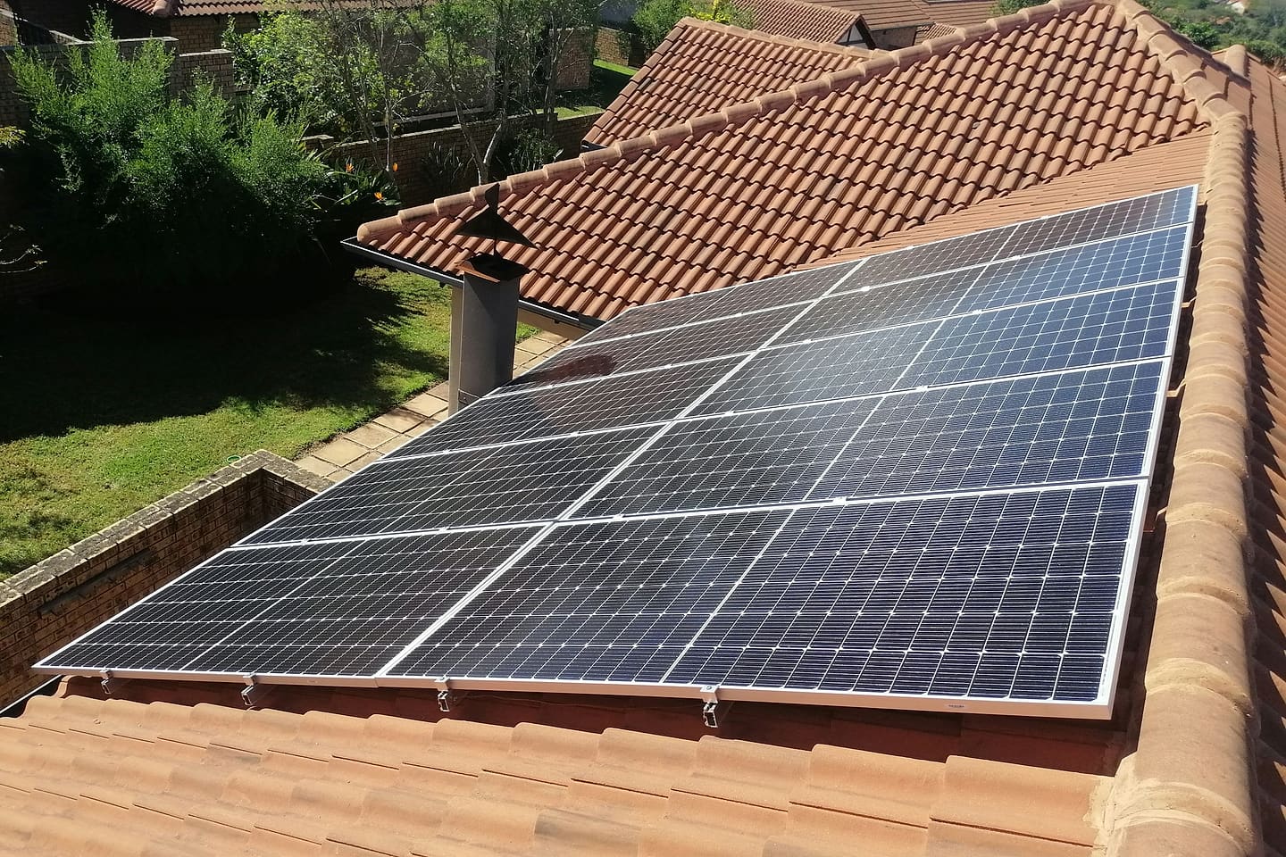 Solar array of dark blue modules installed on a house's red clay-tile roof, with a chimney on the left and the roof's second plane behind it.